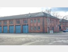 Backsteingebäude mit mehreren blauen Toren, vergitterten Fenstern und einem Satteldach, gelegen an einer Straßenecke. An der Fassade ist eine Leiter angebracht, und ein Schild mit der Aufschrift „Hyflexar“ weist auf einen Hydraulik-Dienst hin.