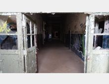 Verlassener Korridor in einem heruntergekommenen Gebäude mit beschädigten Türen, abblätterndem Lack und Graffiti an den Wänden; der Boden ist staubig, die Wände bestehen teilweise aus blauen Fliesen.