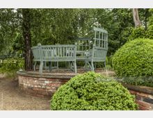 Runde Sitzgruppe aus grün gestrichenem Holz auf einem erhöhten gemauerten Podest in einem Garten, umgeben von Bäumen und gepflegten Sträuchern. Im Hintergrund sind dichte grüne Blätter und ein Kiesweg zu sehen.