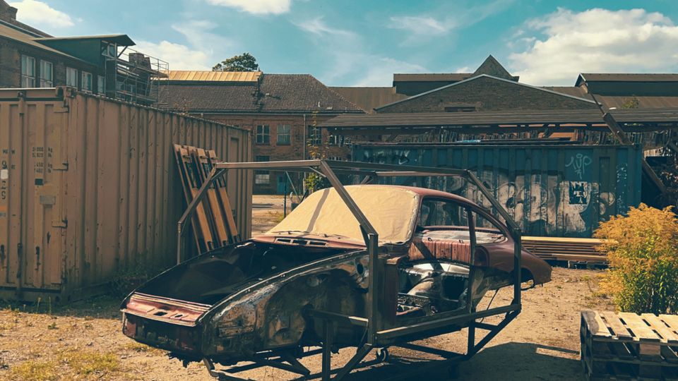 Industriehof mit Schotterboden, umgeben von Containern und Lagerhallen, teils mit Graffiti. Im Vordergrund steht ein ausgebranntes Autowrack in einem Metallrahmen neben Holzpaletten unter blauem Himmel mit Wolken.
