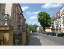 Straßenszene mit Wohnhäusern im historischen Stil, darunter ein rosa Gebäude mit verzierten Balkonen und ein weißes Gebäude mit roten Dachelementen. Die Straße ist von Bäumen gesäumt, daneben sind geparkte Autos und ein Gehweg mit einem niedrigen Ziegelzaun.