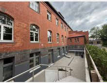 Ehemaliges Backsteingebäude mit modernen Fenstern und einem angrenzenden gepflasterten Innenhof. Im unteren Bereich des Gebäudes sind graue Fassadenelemente, während der Hof mit Gartenmöbeln ausgestattet ist; im Hintergrund sind Bäume und weitere Gebäude erkennbar.