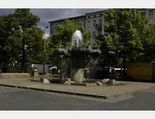 Ein Brunnen auf einem gepflasterten Platz, umgeben von Bäumen und mehrstöckigen Gebäuden im Hintergrund. Der Brunnen hat eine zentrale Wassersäule, die von mehreren Wasserspeiern umgeben ist.