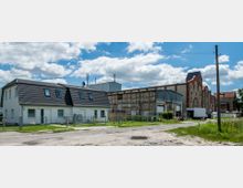 Zweistöckiges Wohnhaus mit dunklem Satteldach neben einer alten, teils verfallenen Industriehalle aus Backstein und Holz. Im Vordergrund befinden sich eine unbefestigte Straße, ein Zaun und Grünflächen, dahinter eine helle, teils bewölkte Himmelkulisse.