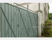 Grünes Eisentor mit diagonalen Verstrebungen und dekorativen Verzierungen in der Mitte, neben einem beigen Gebäude mit Rundbogenfenstern und umgeben von Büschen.