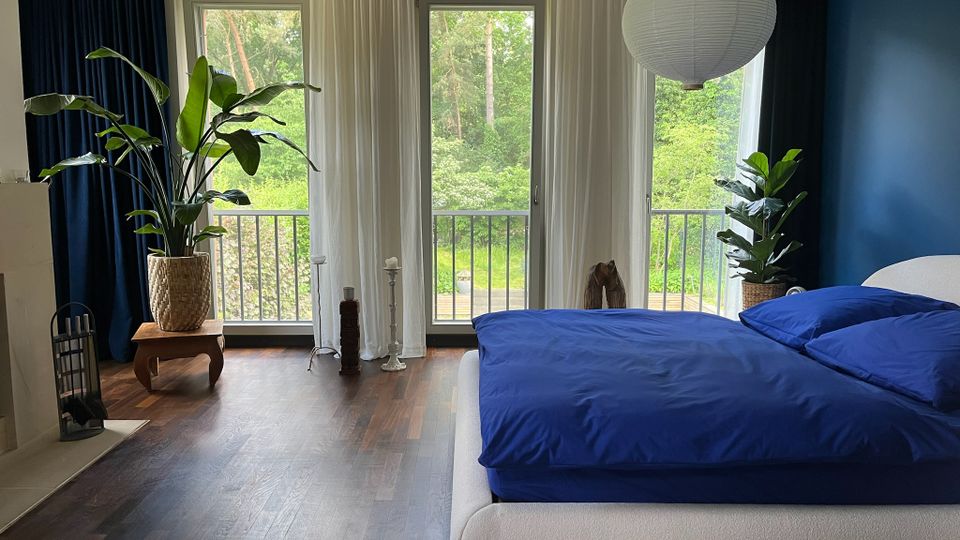 Modernes Schlafzimmer mit dunklem Holzboden, blau gestrichener Akzentwand und großem Bett mit blauer Bettwäsche. Drei bodentiefe Fenster mit weißen Vorhängen öffnen den Blick auf einen Balkon und viel Grün; große Zimmerpflanzen und eine runde Papier-Hängelampe ergänzen die Einrichtung.