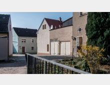 Wohngebiet mit mehreren Einfamilienhäusern, teils mit Putzfassaden in hellen Farben. Im Vordergrund ein kleiner Garten mit blühenden Sträuchern und einem Metallzaun, dahinter ein gepflasterter Weg.