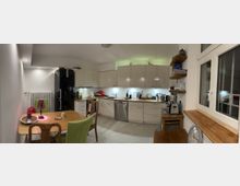 Moderner Küchenraum mit weißen Einbauschränken, einem schwarzen Kühlschrank, einer Spülmaschine und einer Arbeitsfläche mit Küchengeräten. Im Vordergrund steht ein Esstisch mit farblich gemischten Stühlen, daneben befindet sich ein großes Fenster mit sichtbaren Holzfensterbänken und Regalen an der rechten Wand.