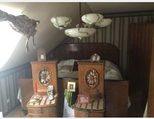 Dachzimmer mit einem antiken Doppelbett aus Holz, flankiert von zwei Nachttischen mit dekorativen Details und persönlichen Gegenständen wie Fotos, einer Kerze und einem Wecker. Die Wände sind mit vertikal gestreifter Tapete gestaltet, und ein Kronleuchter mit Glaslampen hängt von der Zimmerdecke.