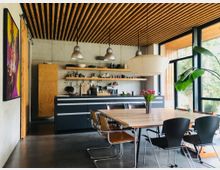 Modern eingerichtete Küche mit offenem Essbereich, einem Holztisch und gemischten Stühlen. Sichtbetonwände, eine Strebenholzdecke, große Fenster mit Blick ins Grüne und eine zentrale Pendelleuchte prägen den Raum.