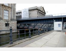 Eine Brücke aus Metallträgern, die über einen Wasserkanal führt, flankiert von historischen Gebäuden aus Stein. Im Vordergrund befindet sich eine mit Dekorationen verzierte Geländerabsperrung; rechts steht ein mobiler weißer Container.