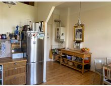 Küche mit hellen Wänden und Holzboden, ausgestattet mit einem großen Edelstahlkühlschrank, einer hölzernen Küchenzeile mit Körben und Küchenutensilien sowie einem Esstisch mit Stühlen. An der Wand hängen ein gerahmtes Bild und eine Gastherme.