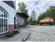 Ein gepflasterter Hof mit einer weiß-roten Fassade eines Gebäudes links, das große Bogenfenster und Holztüren aufweist. Rechts im Bild steht eine alte, orange-gelbe Lokomotive auf einem begrünten Bereich, umgeben von Bäumen.