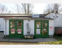 Ein einstöckiges Gebäude mit weiß verputzter Fassade, grünen Doppeltüren aus Glas und einem Schild mit der Aufschrift „fabrik“ auf dem Dachvorsprung. Im Vordergrund befindet sich ein Asphaltweg, im Hintergrund kahle Bäume.