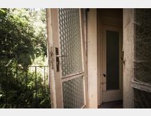 Offene Balkontür mit strukturiertem Glas in einem alten Gebäude, dahinter dichter Bewuchs und ein Geländer, das den Balkon begrenzt. Im Hintergrund ist ein weiterer Innenbereich mit einer geschlossenen Tür sichtbar.