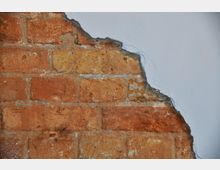 Eine Nahaufnahme einer roten Ziegelmauer, die teilweise von glatter, weißer Putzfarbe überdeckt ist. Der Putz ist stellenweise abgeplatzt und zeigt die darunterliegende Ziegelstruktur.