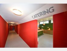 Langer Flur mit roten Wänden und weißem Boden, rechts ein Durchgang zu einem Catering-Bereich mit grüner Wand, Holztheke, weißen Orchideen und dekorativer Beleuchtung. An der Decke hängt ein Schild mit der Aufschrift "Catering".