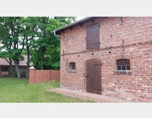 Backsteingebäude mit braunen Holztüren und kleinen Fenstern mit Ziegelbögen, daneben eine niedrige Ziegelmauer. Im Hintergrund stehen Bäume und ein weiteres Gebäude mit einem Ziegeldach.