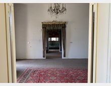 Blick in einen weitgehend leeren Raum eines historischen Gebäudes mit weiß verputzten Wänden, Kronleuchter und einem roten Orientteppich im Vordergrund. In der Mitte führt eine mit schweren Vorhängen und goldener Zierblende gerahmte Türöffnung in weitere Räume mit Fenstern.