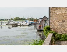 Ein kleiner Hafen mit mehreren festgemachten Motorbooten und einem Hausboot am Ufer. Im Vordergrund ein Teil einer Backsteinmauer und Pflanzen, im Hintergrund Bäume und Stromleitungen.