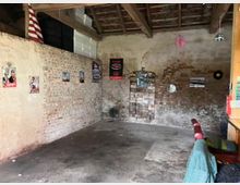 Ein rustikaler Raum mit unverputzten Backsteinwänden, einem Sichtdach mit Holzbalken und Betonboden. An den Wänden hängen Retro-Schilder, Poster und einzelne Deko-Elemente wie Schallplatten; im rechten Vordergrund ist ein Teil eines gepolsterten Stuhls und einer Holztheke sichtbar.