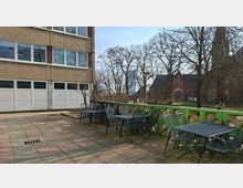 Außenbereich mit gepflasterter Terrasse neben einem mehrstöckigen Gebäude mit großen Fensterreihen und mehreren weißen Garagentoren. Entlang einer niedrigen grün gestrichenen Betonbrüstung stehen Metalltische und -stühle; im Hintergrund sind kahle Bäume, eine rote Backsteinkirche und weitere Gebäude zu sehen.