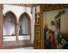 Innenraum einer Kirche mit roten Ziegelbögen, dahinter sichtbare Orgelpfeifen. Rechts ein detailreiches, vergoldetes Altarbild mit der Darstellung der Kreuzigung Jesu.