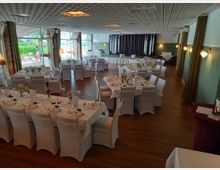 Ein großer, elegant dekorierter Festsaal mit weiß eingedeckten Tischen und Stühlen, Blumengestecken und gedecktem Geschirr. Im Hintergrund sind große Fenster mit Blick nach draußen sowie eine Bühne und Vorhänge sichtbar.