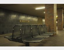 Untergrundbahnhof mit dunkelblauen Wandfliesen, einer Sitzbank aus Metallgitter und roten Backsteinsäulen. An der Wand ist ein Schild mit der Aufschrift „Westphalweg“ angebracht.