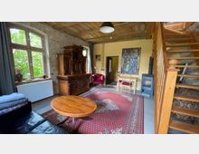 Wohnzimmer in einem rustikalen Stil mit unverputzten Ziegelwänden, freiliegenden Holzbalken an der Decke und einer Holztreppe. Der Raum enthält ein großes Holzbuffet, einen runden Holztisch, einen roten Teppich, eine kleine Sitzecke mit einem roten Sessel und einen Kaminofen an der Wand.