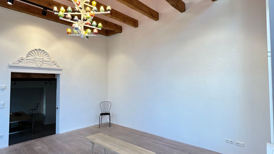 Heller, nahezu leerer Innenraum mit breiten Holzdielen, weiß verputzten Wänden und sichtbaren Holzbalken unter einer roten Deckenfläche. Ein farbiger Kronleuchter hängt von der Decke, dazu gibt es eine kleine Bank in der Mitte und einen Durchgang mit dekorierter Stuckumrahmung links.