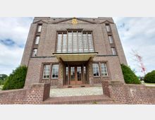 Backstein-Gebäude mit symmetrischer Fassade, großen Fenstern und einem Eingangsbereich mit Holztüren und einem überstehenden Vordach. Der umliegende Bereich besteht aus gepflastertem Boden und niedrigen Backsteinmauern, umgeben von Sträuchern.