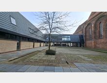Innenhof zwischen einem modernen, dunkel verkleideten Gebäude und einer großen Backsteinkirche mit hohen Rundbogenfenstern. Der gepflasterte Platz hat breite Steinwege, niedrige Stufen und einen zentralen, blattlosen Baum vor einer Glasfassade am Hofende.
