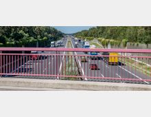 Eine Autobahn mit mehreren Fahrspuren, aufgenommen von einer Brücke mit rot lackiertem Geländer. Beidseitig der Fahrbahn befinden sich Schallschutzwände, teils mit Pflanzen bewachsen, und die Umgebung ist von Wald und Grünflächen gesäumt.