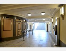 Eingangsbereich einer historischen U-Bahn-Station mit hellen Fliesen, runden Deckenleuchten und Wandlampen im klassischen Stil. Links befindet sich eine geschwungene Wandverkleidung mit Holzdetails, rechts ein Münztelefon, und geradeaus führen Treppenstufen nach draußen.