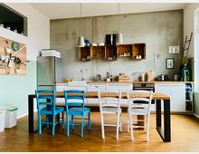 Helle, modern gestaltete Wohnküche mit weißen Unterschränken und einer Holzarbeitsplatte. An zentraler Stelle steht ein Esstisch mit blauen und weißen Stühlen, darüber hängen Hängeleuchten; an der Wand befinden sich Regale aus Holzkisten.