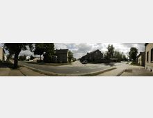 Straßenszene in einem Wohngebiet mit älteren Gebäuden, teils aus Ziegel- oder Putzfassaden, umgeben von Bäumen und Gehwegen. Eine Kreuzung ist sichtbar, mit parkenden Autos und leicht bewölktem Himmel im Hintergrund.