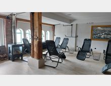 Innenraum eines gemütlichen Ruhebereichs mit Liegestühlen, einem Ofen mit Holzscheiten und freiliegenden Holzbalken. Die Wände sind weiß verputzt, teils mit Ziegeln, und einige Fenster mit grünen Rahmen sorgen für Tageslicht.