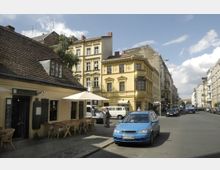 Straßenszene in einer Stadt mit historischen Gebäuden, darunter ein kleines gelbes Haus mit Ziegeldach und Außenbestuhlung vor einer Weinbar. Im Hintergrund stehen mehrstöckige Altbauten mit verzierten Fassaden, und auf der Straße parken Autos.