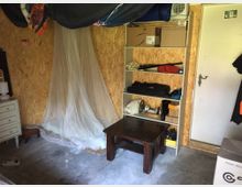 Ein einfach möblierter Raum mit Holzwänden und Betonboden, ausgestattet mit einem niedrigen Holztisch, einem offenen Regal mit Kartons und Gegenständen sowie einem kleinen Bett mit einem hängenden Moskitonetz. Rechts befindet sich eine weiße Tür, links ein kleines Schränkchen.