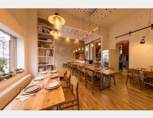 Ein gemütlicher Speiseraum mit Holztischen und -stühlen, gedeckt mit Tellern und Gläsern, beleuchtet von hängenden Vintage-Glühbirnen. Die Wände sind aus Backstein und weiß gestrichenem Putz, mit einem Regal voller Bücher und Dekorationen sowie einer großen Fensterfront links.