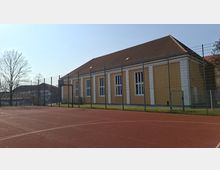 Eingezäunter Sportplatz mit rotem Belag vor einem langen, gelb-weißen Gebäude mit Satteldach und hohen, schmalen Fenstern. Im Hintergrund stehen weitere Gebäude und kahle Bäume unter klarem Himmel.