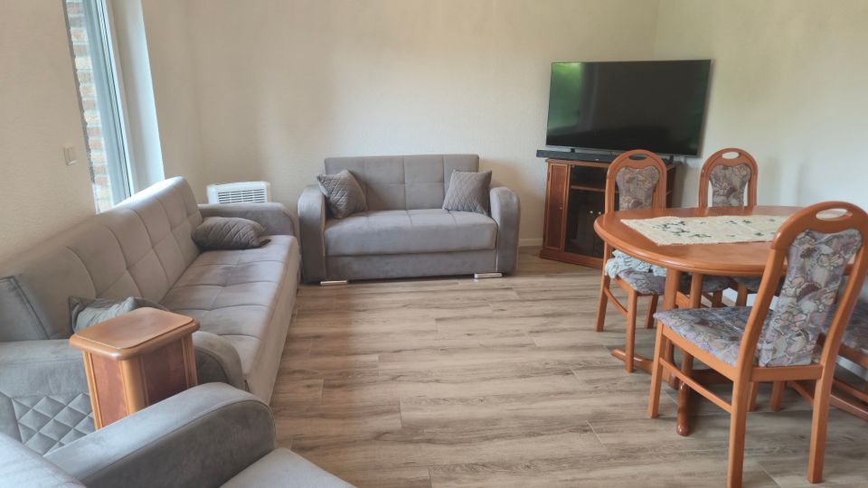 Helles Wohn- und Esszimmer mit zwei grauen Sofas, Holzboden und großem Fenster an der linken Wand. Rechts stehen ein runder Holzesstisch mit gepolsterten Stühlen sowie ein Fernseher auf einem Eckschrank.