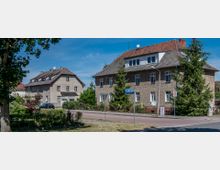 Straßenszene mit zwei zweistöckigen Wohnhäusern im traditionellen Baustil, mit verputzten Fassaden und roten Ziegeldächern. Umgeben von Bäumen, kleinen Gärten und Gehwegen, befindet sich ein Straßenschild im Vordergrund sowie Verkehrszeichen an der Ecke einer ruhigen Straße.