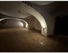 Großer, leerer Keller- oder Gewölberaum mit weiß verputzten Backsteinbögen und niedrigem Tonnengewölbe. Holzplankenboden und wenige Wandlampen beleuchten die gebogenen Durchgänge und Nischen.