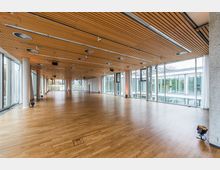 Großer, moderner Veranstaltungsraum mit Holzfußboden und einer Decke aus Holzpaneelen, die mit eingelassenen Leuchten und Lüftungselementen ausgestattet ist. Die Wände sind größtenteils aus Glas, wodurch viel Tageslicht hereinfällt, und bieten Blick auf die umliegenden Gebäude und Bäume.