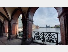 Blick durch Ziegelbogen einer historischen Brücke mit verziertem Geländer auf einen breiten Fluss, umgeben von modernen und alten Gebäuden, unter einem leicht bewölkten Himmel.