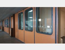 Flur in einem Gebäude mit orangefarbenen Wänden, in die große Fenster mit dunklen Rahmen eingelassen sind; im Inneren sind helle Deckenleuchten und ein Treppengeländer sichtbar. Der Boden ist aus Beton, und eine offene Tür führt in einen angrenzenden Raum.