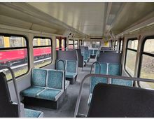 Innenansicht eines leeren Regionalzugwagens mit gepolsterten Sitzen in grünen Mustern, langen Fensterreihen und Gepäckablagen unter der Decke. Draußen sind ein roter Zug und ein Bahnsteig sichtbar.