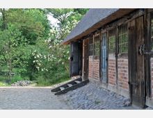 Historisches Gebäude mit roten Ziegelwänden, Holztüren und Reetdach, umgeben von blühenden Sträuchern und Bäumen. Der gepflasterte Weg führt an der rechten Gebäudeseite entlang durch eine ländliche, grüne Umgebung.